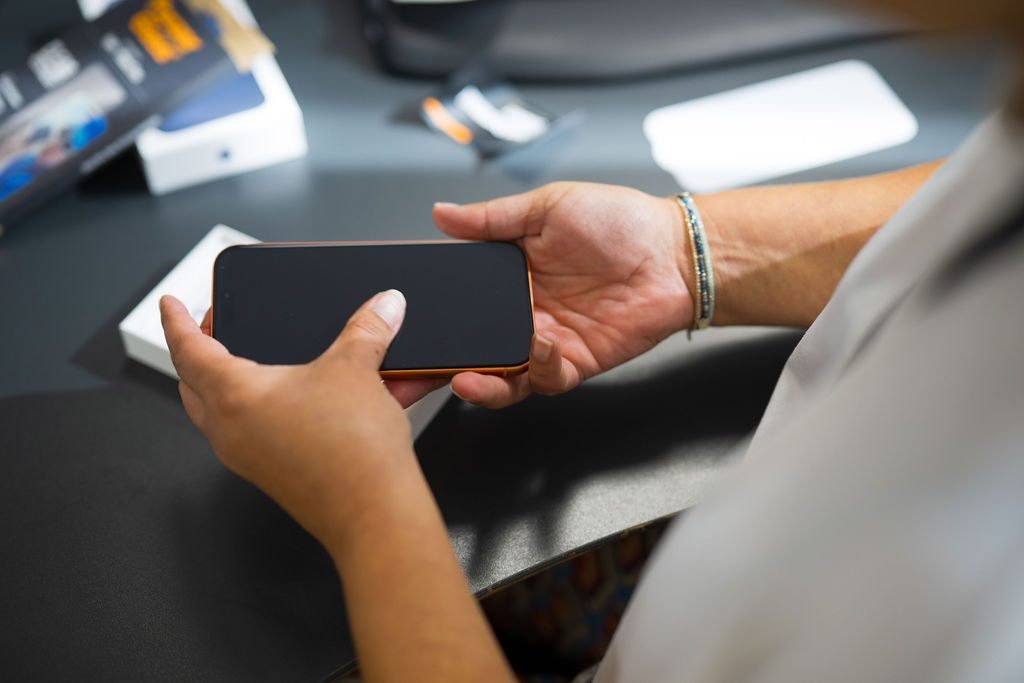 Et nærbillede af en persons hænder, der holder en sort smartphone horisontalt over en mørk bordplade. Personen er i gang med at pakke telefonen ud eller montere beskyttelse, og i baggrunden ses produktæsker og emballage.