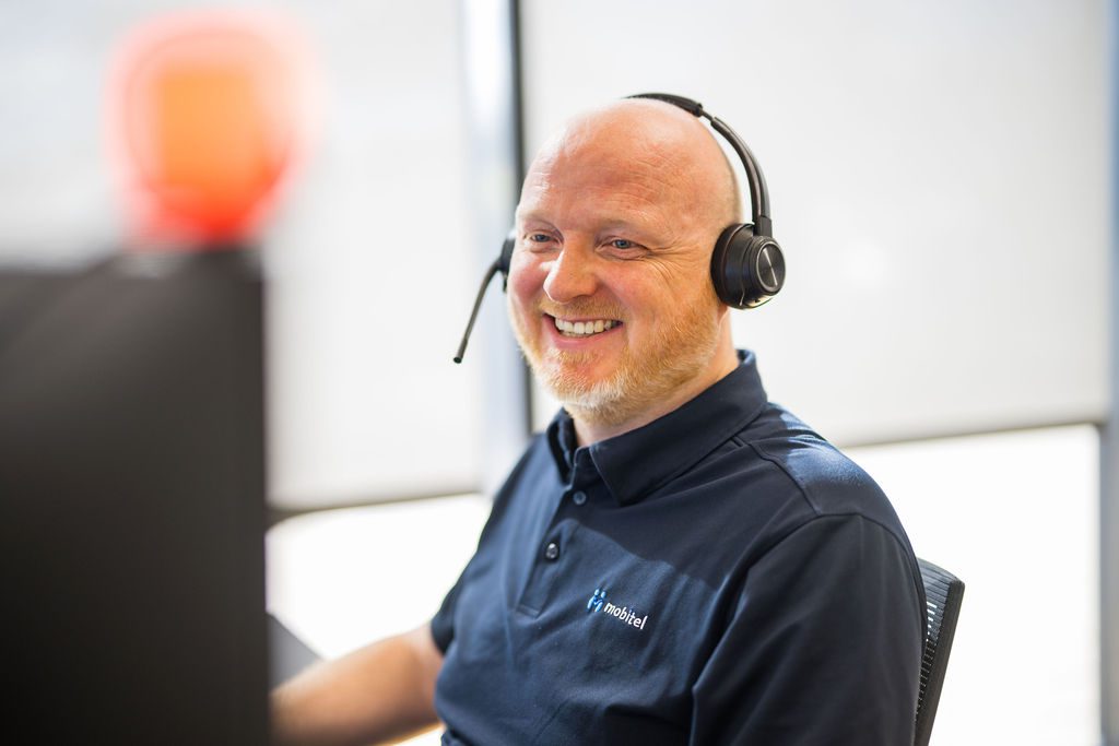 En smilende, skaldet mand med kort lyst skæg sidder ved en computer og bærer et professionelt headset. Han er iført en mørkeblå polo-shirt med et hvidt "mobitel" logo.